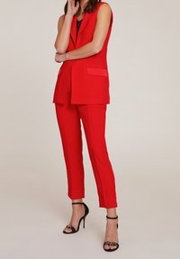 Röd skräddarsydd kostym med ärmlös kavaj, som har två fickor, slätt tyg och smala byxor. Bärs med svarta högklackade sandaler.