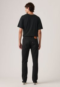 Μαύρο κοντομάνικο μπλουζάκι, συνδυασμένο με μαύρα τζιν τζιν με δερμάτινο έμπλαστρο και χαρακτηριστικές πίσω τσέπες, φορεμένο με μαύρα αθλητικά παπούτσια.