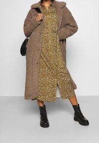 Brązowy pluszowy płaszcz z kołnierzem, zapinany na guziki, nałożony na musztardową sukienkę w kwiaty do kolan. Całość dopełniają czarne, sznurowane botki.