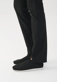 Svarte slip-on sko med teksturert overdel og hvit gummisåle, plassert ved siden av skreddersydde svarte bukser, som viser et minimalistisk design.
