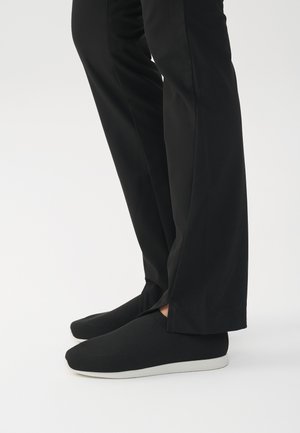 Schwarze Slipper mit einer strukturierten Oberfläche und weißer Gummisohle, zusammen mit maßgeschneiderten schwarzen Hosen präsentiert, die ein minimalistisches Design aufweisen.