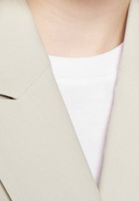 Ljus beige kavaj med skarpa slag, lager på lager över en vit crew neck-topp. Tyget verkar slätt med en lätt glans.