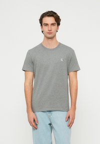 Szürke pamut póló kerek nyakkal, rövid ujjakkal és egy kis fehér logóval a mellkason, világoskék farmernadrággal bemutatva.