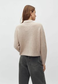 Világos rózsaszínű, kötött pulóver rövidített fazonban, sálgallérral és finom, gyémánt alakú kötésmintákkal az ujjain.