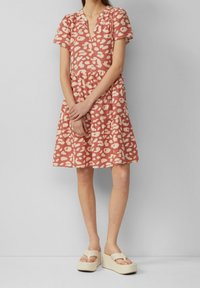 Donna che indossa un vestito a maniche corte, di colore corallo rosa, con un motivo e scollatura a V, in piedi con le mani congiunte e indossando sandali con zeppa beige.