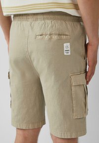 Beige Cargo-Shorts aus strukturiertem Stoff, mit elastischem Bund, Seitentaschen und einem Etikett auf der Rückseite.