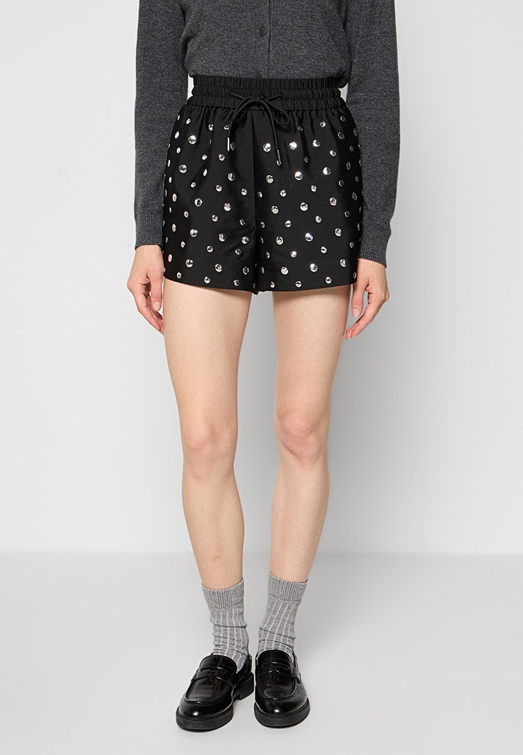 Schwarze Shorts mit einem elastischen Bund, Kordelzug und verstreuten silbernen Nieten. Getragen mit grauen Socken und dunklen Schuhen.