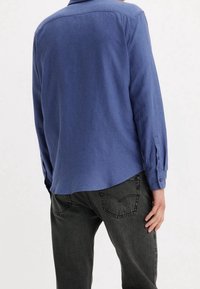 Niebieska koszula z długim rękawem wykonana z miękkiej tkaniny, ze mankietem zapinanym na guziki i lekko zaokrąglonym dołem, zestawiona z ciemnymi dżinsami.