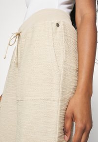 Zbliżenie na osobę w beżowych, texturednych spodniach dresowych z elastycznym pasem na sznurek oraz małym metalowym guzikiem przy kieszeni.