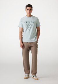 Tricou din bumbac de culoare albastru deschis, cu un design grafic triangular în albastru mai închis, asortat cu pantaloni bej, largi, și adidași multicolori.