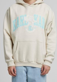 Beige hoodie med en stor blå grafisk tryck av en fågel och texten "KARL KAN" på framsidan. Inkluderar en framficka och ribbade  ärmslut.