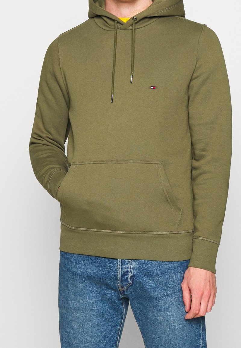 Felpa verde oliva con cappuccio e cordini, tasca a marsupio, polsini e orlo a coste. Piccolo logo sul petto. Indossata con jeans blu.