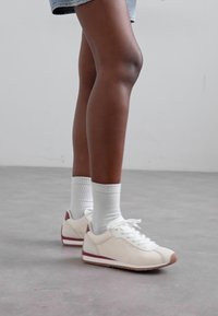 Sneakers bianchi con accenti bordeaux, materiale simile alla pelle, profilo basso, punta arrotondata, stringhe e suola in gomma beige testurizzata, abbinate a calzini bianchi alla caviglia.