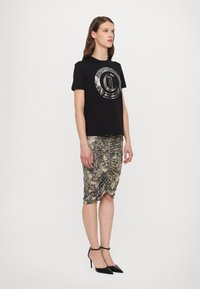 T-shirt noir avec un design graphique métallique, associé à une jupe froncée en imprimé serpent beige et noir. Le modèle porte des talons aiguilles noirs.