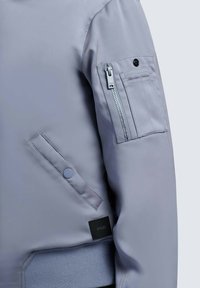 Manche de veste gris clair avec une poche zippée à rabat boutonné et des surpiqûres sur le haut du bras, ainsi qu'une poche latérale avec bouton-pression.