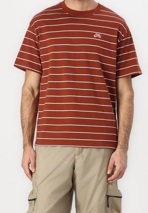 Homme portant un T-shirt à manches courtes couleur rouille-orange avec des rayures horizontales blanches et un short cargo beige sur un fond neutre.