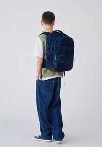 Marineblaue Rucksack mit geometrischen Mustern und grünen Akzenten, ausgestattet mit mehreren Fächern und gepolsterten Trägern, getragen von einem Kind.