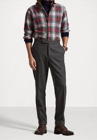 Camicia con bottoni a quadri rossi e verdi, maniche arrotolate, abbinata a pantaloni grigi testurizzati e scarpe di pelle marrone.