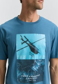 Mėlyna medvilninė marškinėliai su grafiniu spaudiniu, vaizduojančiu sraigtasparnį mėlyname fone. Tekstas sako "TOM TAILOR" ir "TEAM AERONAUT CAPACITY."
