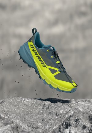 Scarpa da trail running Dynafit blu e giallo neon sospesa sopra un terreno roccioso con particelle di terra che fluttuano intorno.