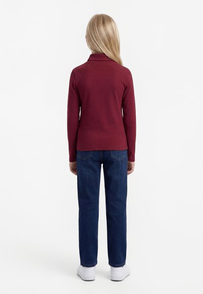 Kind met lang blond haar met de rug naar de camera, draagt een bordeauxrode top met lange mouwen, blauwe spijkerbroek en witte sneakers tegen een effen witte achtergrond.
