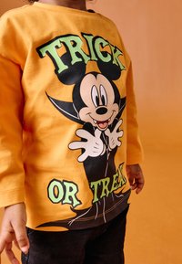 Gul langærmet skjorte med stor grafik af en tegneseriefigur og teksten "TRICK OR TREAT" i grønt, båret med mørke bukser.