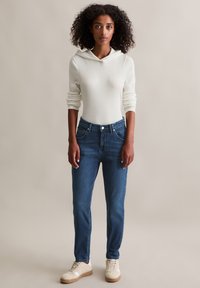 Witte geribbelde lange mouwen top met een capuchon, gecombineerd met donkerblauwe spijkerbroeken en crèmekleurige sneakers met bruine zolen. Eenvoudige en casual outfit.