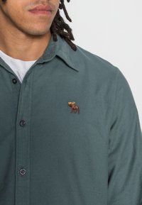 Donkergroen overhemd van een zachte stof met een knoopsluiting, met een klein geborduurd elandlogo in bruin en geel op de borst.