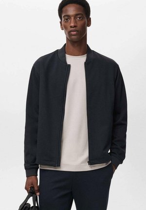 Black bomber jacket with a minimalistic design, featuring a round collar and zip front. Worn over a light-colored crew neck shirt.