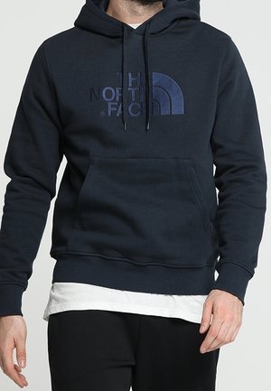 Hombre vestido con una sudadera con capucha azul marino de The North Face con bolsillo frontal tipo canguro y una camisa blanca debajo, de pie con los brazos relajados.