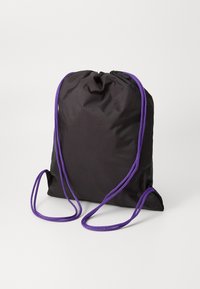 Sac à dos à cordon noir avec des cordons violets, resserré en haut, reposant à la verticale sur une surface blanche unie.