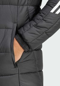 La chaqueta de plumón negra cuenta con un bolsillo con cremallera, puños elásticos, un patrón acolchado horizontal y rayas blancas en la manga como acento.