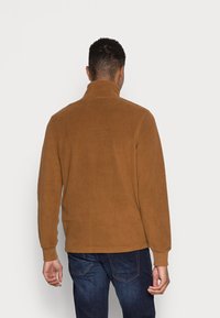 Uomo che indossa una giacca in pile marrone con colletto alto e jeans blu scuro, in piedi di spalle contro uno sfondo chiaro e uniforme.