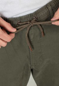 Olijfgroene broek met een elastische tailleband, voorzien van een gevlochten trekkoord met bruine accenten. Close-up van het taillebandgebied.