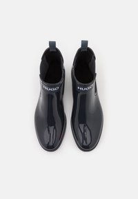 Mustat kiiltävät nilkkurit, joissa joustavat sivut ja vetotappi. Yläosassa näkyvä, valkoinen "HUGO"-logo. Sileä materiaali.