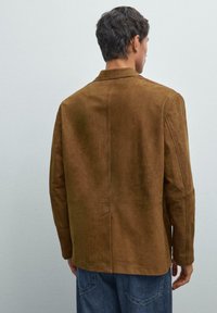Braune Wildlederjacke mit Kragen, die eine glatte Textur und klare Linien zeigt. Sie hat eine zentrale Naht und eine lockere Passform, kombiniert mit Jeans.
