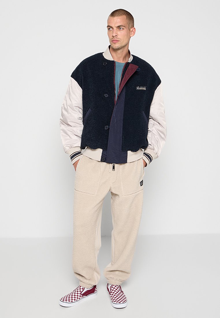 Giacca fuzzy blu navy e crema con chiusura a bottoni, maniche a contrasto, abbinata a pantaloni in pile beige e scarpe slip-on a quadri rossi.