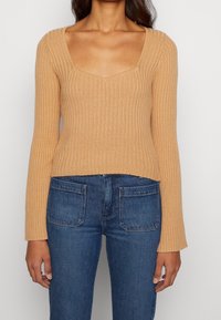 Beige ribbad stickad tröja med fyrkantig halsringning och långa ärmar, med en kortad nederkant och texturerad finish. Bärs med blå jeans.