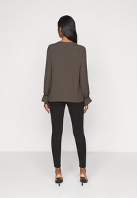 Blusa lunga marrone a maniche lunghe con polsini arricciati, abbinata a jeans skinny neri e tacchi. Tessuto liscio, vestibilità rilassata.