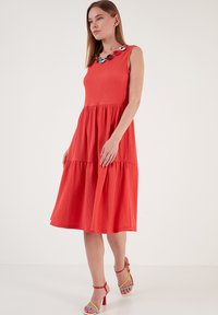 Rotes, gestuftes Midikleid aus strukturiertem Stoff, ärmelloses Design und ein mehrfarbiger, verzierrer Ausschnitt. Kombiniert mit mehrfarbigen, hochhackigen Sandalen.