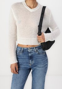 Suéter de punto blanco, recortado, con escote en V y mangas largas, combinado con jeans azules de tiro alto y un bolso negro con una correa con logo.