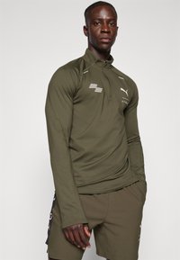 Olijfgroene longsleeve sporttop met ritskraag, uitgevoerd met reflecterende accenten en een logo. Glad, rekbaar materiaal dat is ontworpen voor comfort.