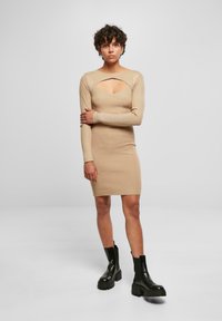 Robe beige à manches longues côtelée avec un décolleté découpé, silhouette ajustée et longueur jusqu'aux genoux. Associée à des bottines noires.