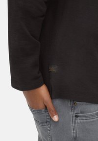 Sort langærmet skjorte med tekstureret stof og sideslidsdetalje; har en lille etiket nær kanten. Hånd hviler i grå denimlomme.