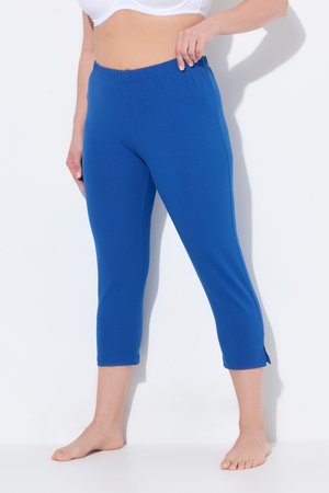 Frau trägt blaue gekürzte Leggings mit elastischem Bund, steht barfuß und zieht den Bund leicht nach außen.