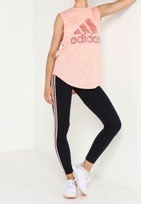 Jasnoróżowy bezrękawnik Adidas z dużym logo, w połączeniu z czarnymi legginsami z różowymi paskami po bokach i białymi butami sportowymi.