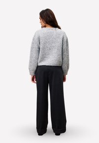 Vrouw met haar rug naar de camera, gekleed in een lichtgrijze, pluizige trui en luchtige zwarte wijde broek tegen een effen witte achtergrond.