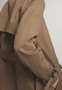 Gros plan d'une personne portant un trench beige avec une ceinture et un poignet boutonné, main glissée dans une poche.