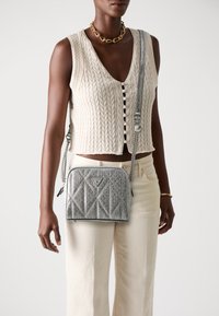 Geantă crossbody gri, capitonată, cu accente de argint și logo. Include o curea din lanț și închidere cu fermoar, având detalii cu textură.