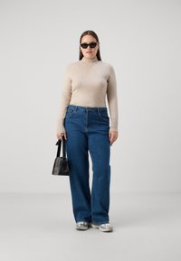 Bež pulover s turtleneck izrezom, modri široki traperji, črne sončne očala, srebrni čevlji in mala črna torbica. Enobarven ozadje.